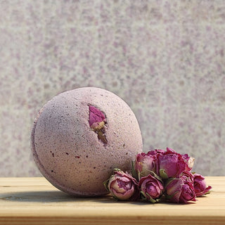 A Bath Bomb Sampler 5 Pack bath bomb, purple with a small flower, sits on wood beside dried pink roses. The soft pastel background complements the relaxing scene.
