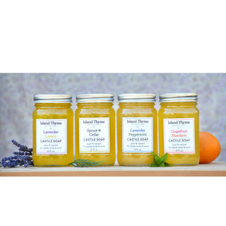 Four jars of Island Thyme Lavender Lemon Castile Soap, made with essential oils and eco-friendly packaging. Each jar features yellow liquid and is surrounded by lavender sprigs and an orange.