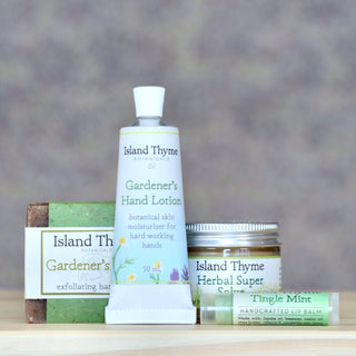 The Gardeners Gift Set from Island Thyme Botanicals features a gardener’s scrub bar, Gardener’s Hand Lotion, Herbal Super Salve jar, and Tingle Mint Lip Balm tube arranged together.