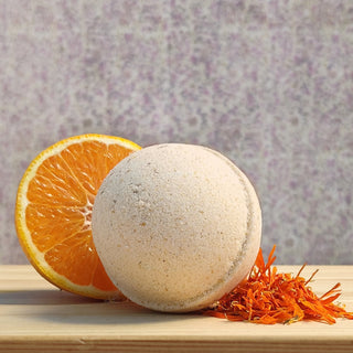 The Sweet Orange Calendula Bath Bomb rests on wood beside sliced orange and orange petals. Made with calendula-infused sweet almond oil, it is set against a softly blurred purple background.